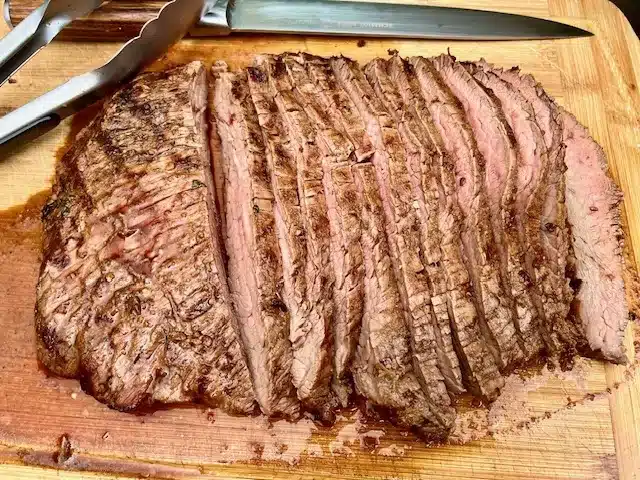 Flank Steak on a cutting board with a knife and tongs.